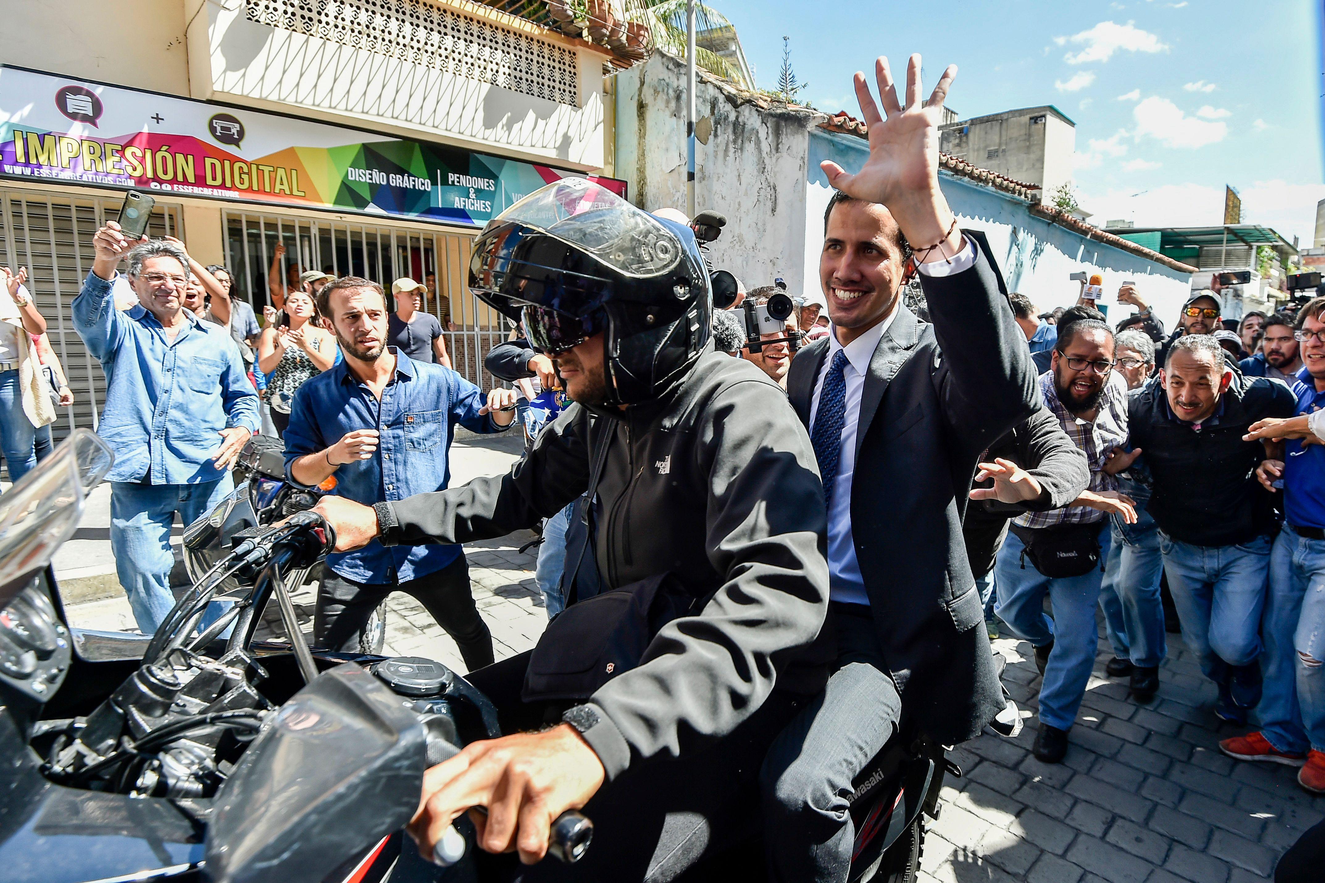 Juan Guaido sits on the back of a motorbike behind a driver. He's waving at a crowd of people.