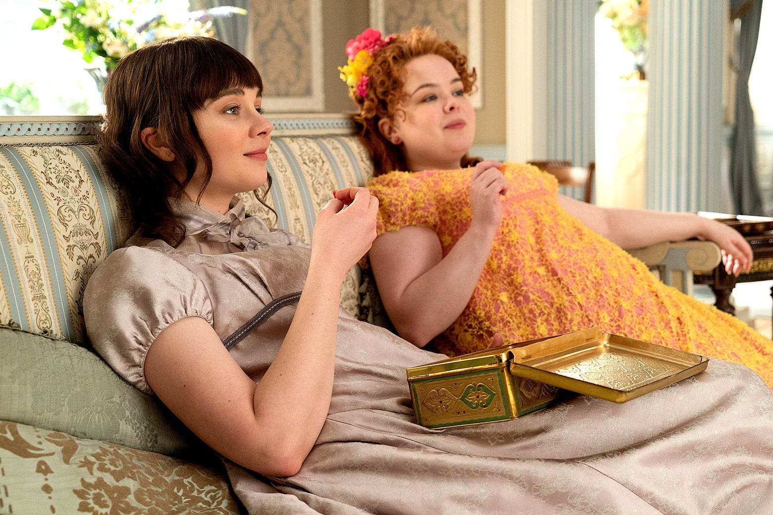 Claudia Jessie and Nicola Coughlan sit slumped on a couch. Jessie wears a high-neck, satiny gown in a muted beige tone; Coughlan wears a busy yellow and orange gown with ruffles.