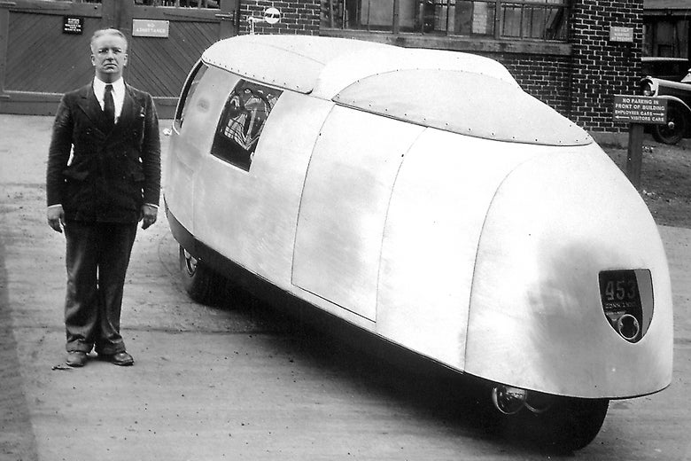 A man stands next to an ovoid car with a smooth exterior. 