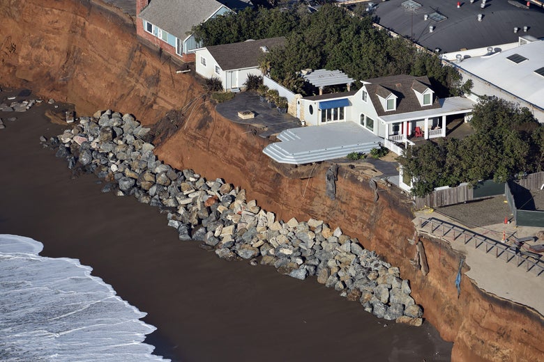 Houses hang just on or over the edge of a cliff above a beach. 