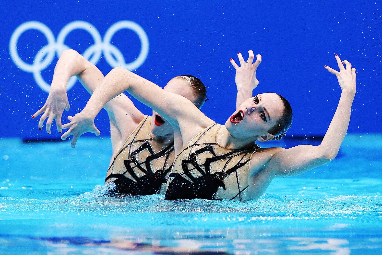Two women, wearing bedazzled suits with giant spiders on them, raise their arms in the air as they swim in a pool.