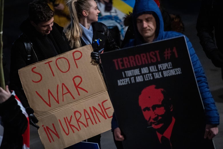 One person holds a sign that says "Stop War in Ukraine" while another holds a sign that says "Terrorist #1: I torture and kill people! Accept it and let's talk business!" over a picture of Putin