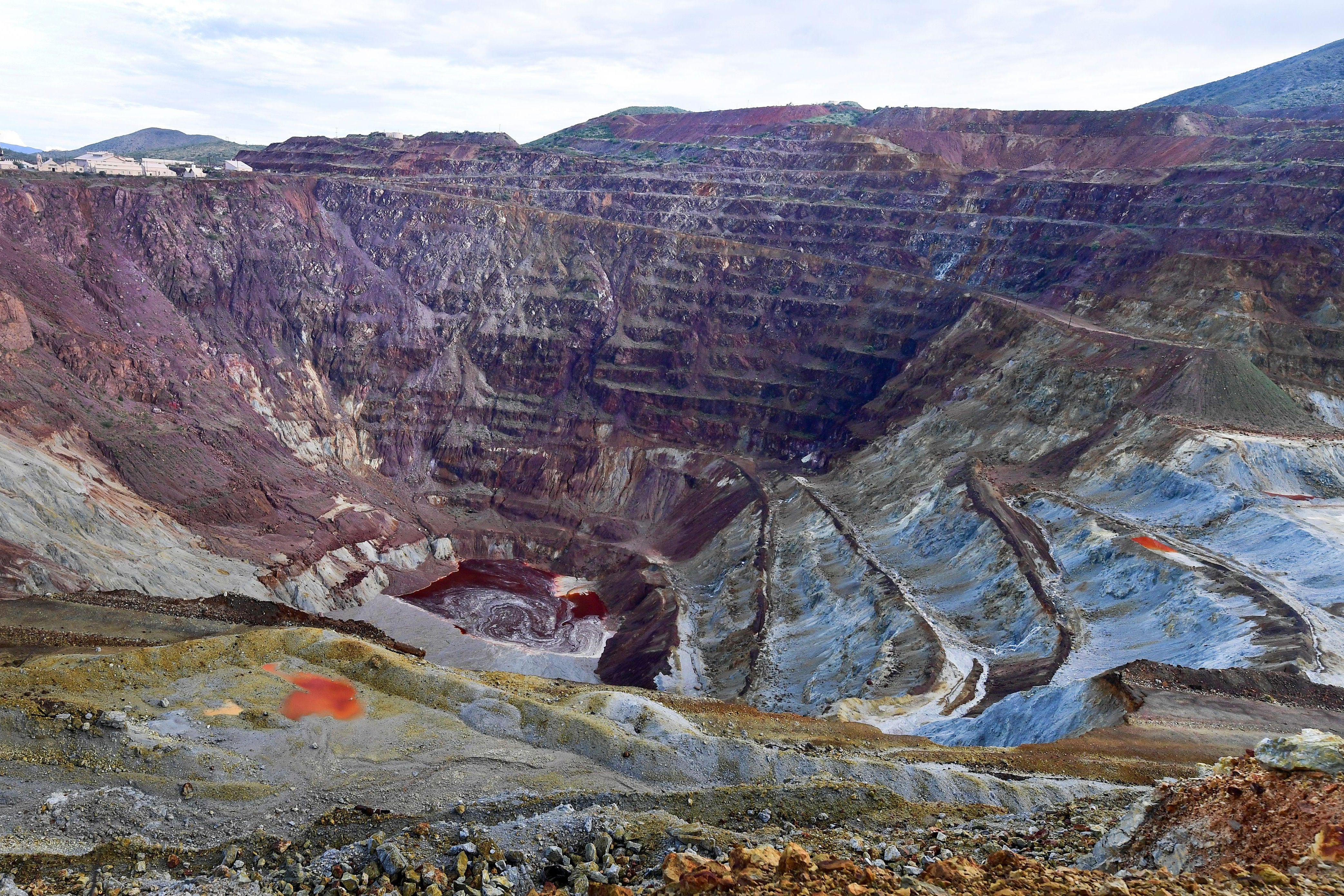 A large open pit mine. The walls surrounding the pit are layered with gray and maroon rock. 