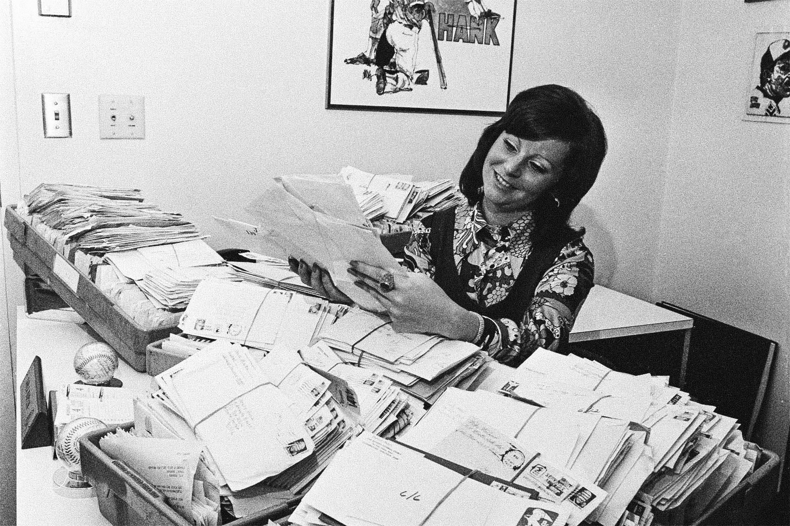 A woman sitting at a desk that's covered with huge piles of unopened letters.