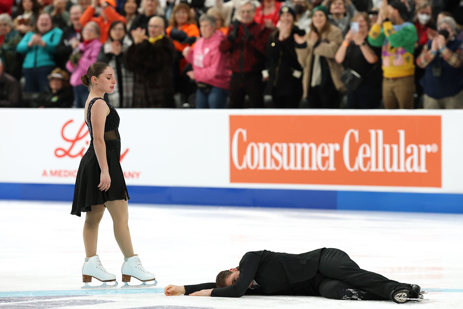 One ice dancer lying on the ice as if dead while the other stands over him. 