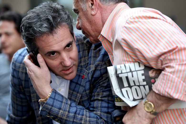 Michael Cohen leans over while holding a cell phone to his right ear while an unidentified man holding a New York Post headline about Donald Trump speaks into his other ear.