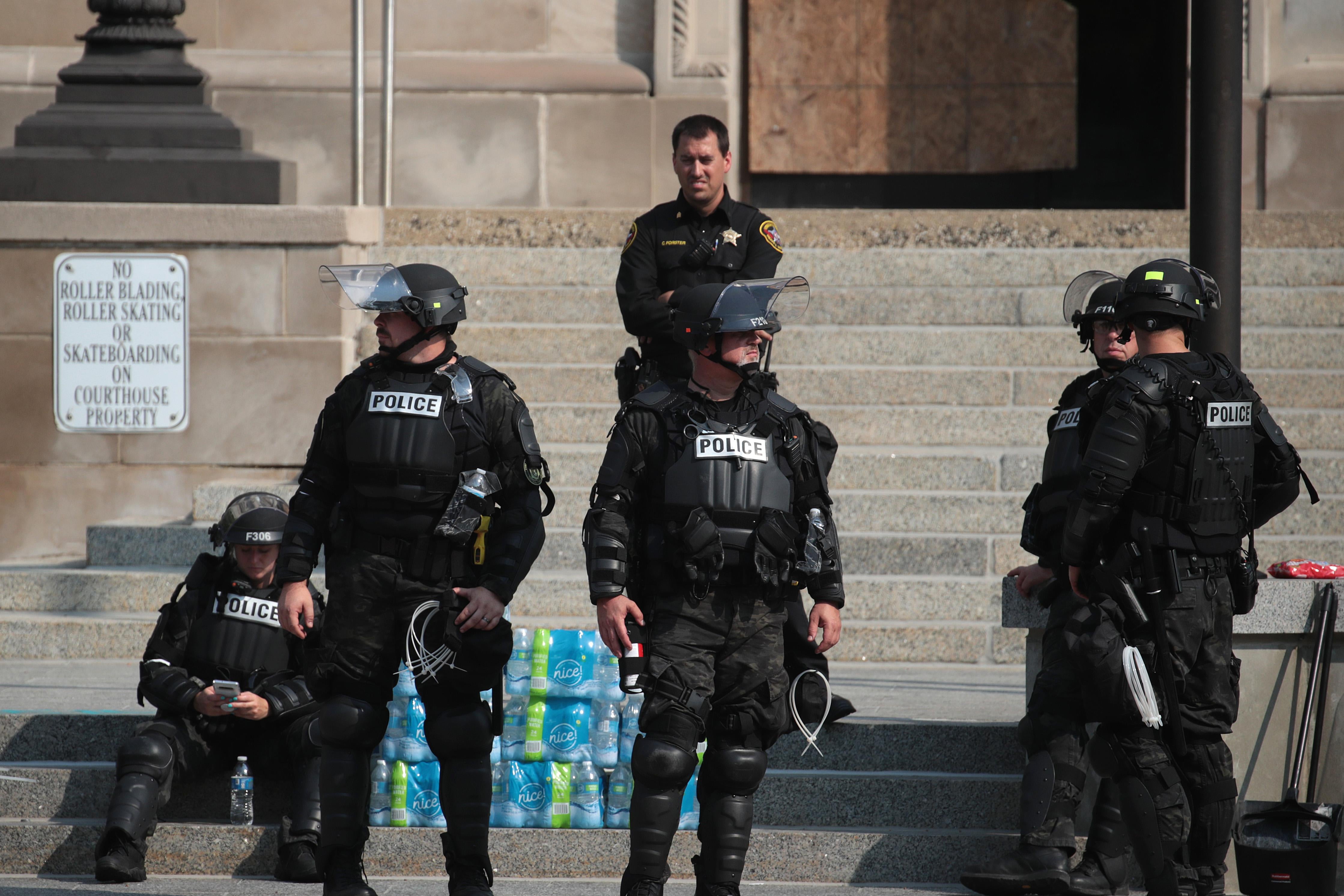 Six police officers dressed in riot gear stand in front of a courthouse. 