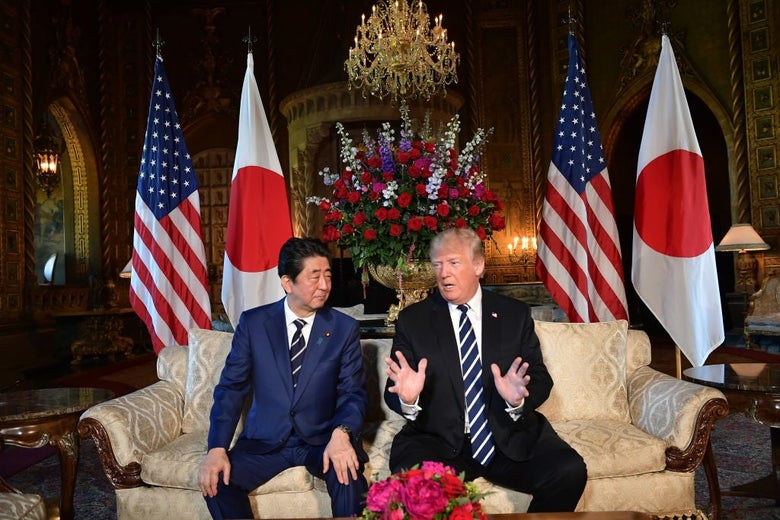 Trump gestures while Abe listens as the two are seated on a cream-colored couch in an ornate living room. Two American and two Japanese flags are behind the men.