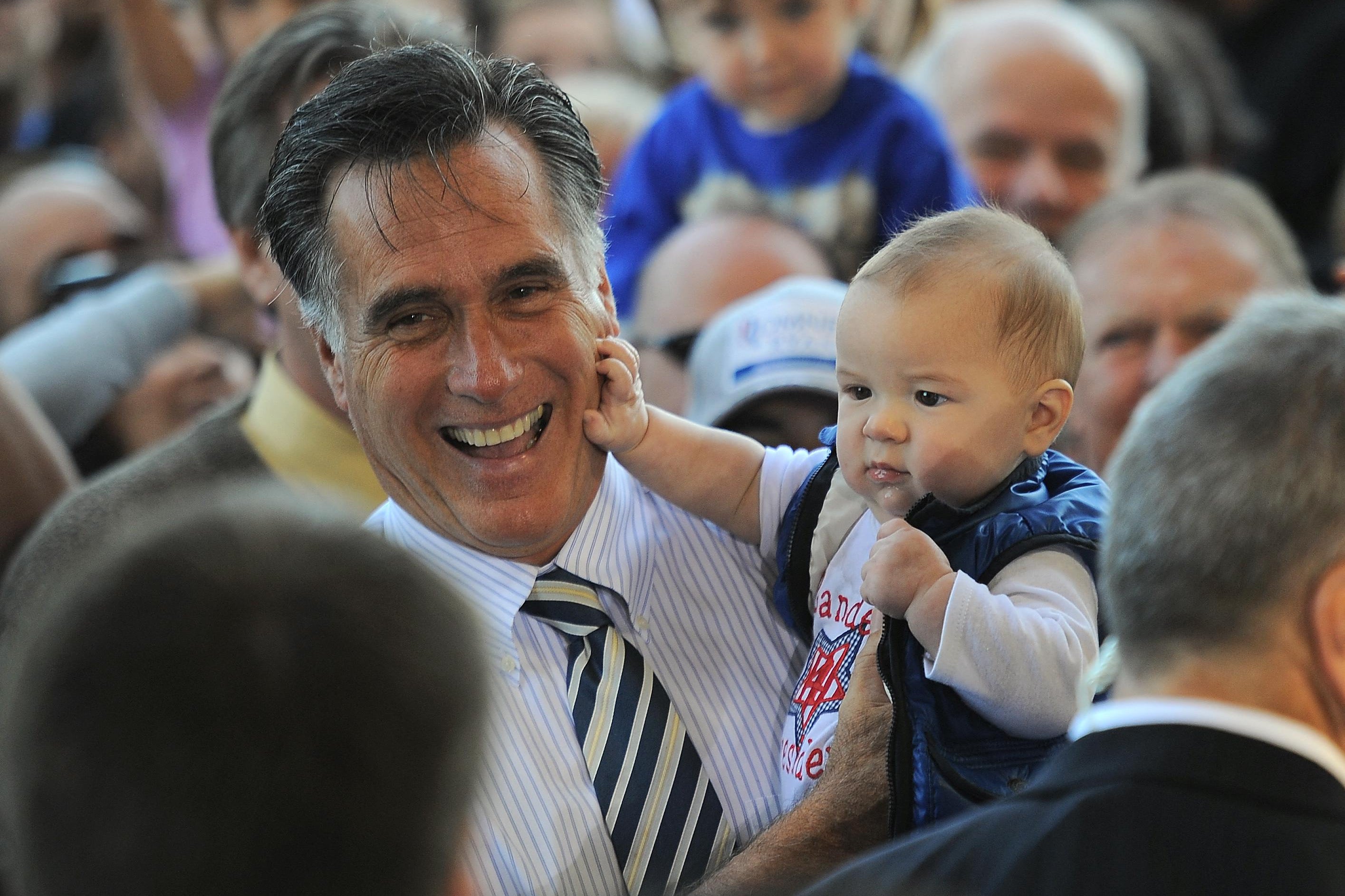 Mitt Romney laughs as he holds a baby in a crowd of people. The baby is grabbing Romney's cheek.