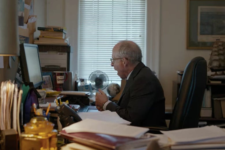 Elliot looks at a computer while sitting at a desk with lots of papers and folders on it.