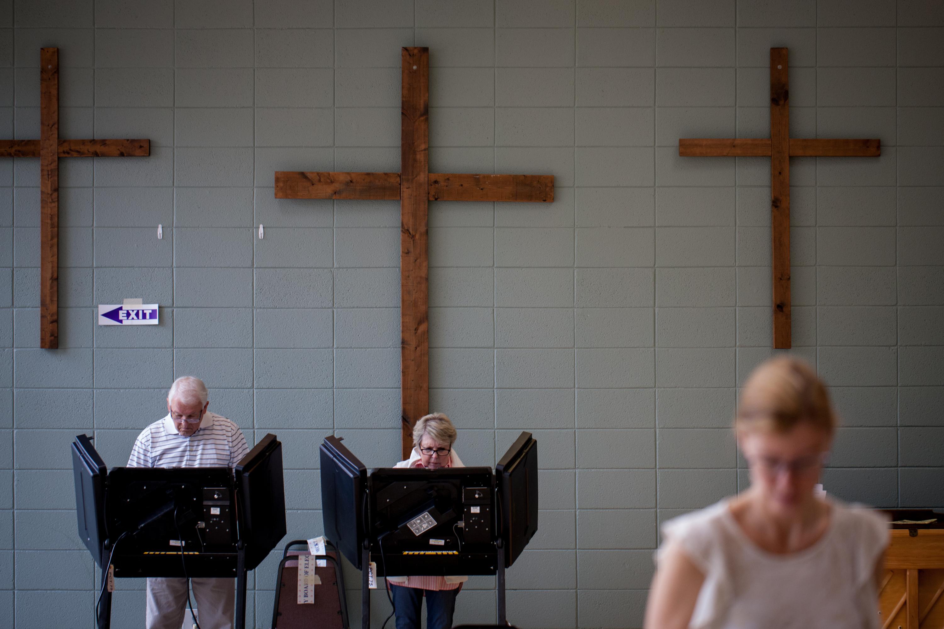 Two people at voting booths. Three Christian crosses hang on the wall behind them.