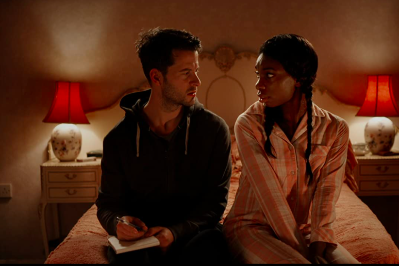 Robert Lonsdale, holding a notepad and pen, sits on the edge of a bed with Michaela Coel.