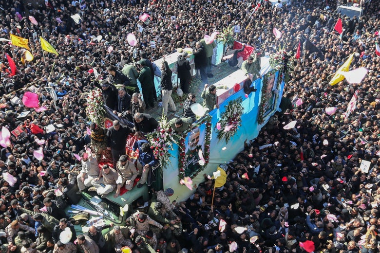 A large crowd surrounds a truck with people on top of it. The truck features a photo of Qassem Soleimani framed in flowers on the side.