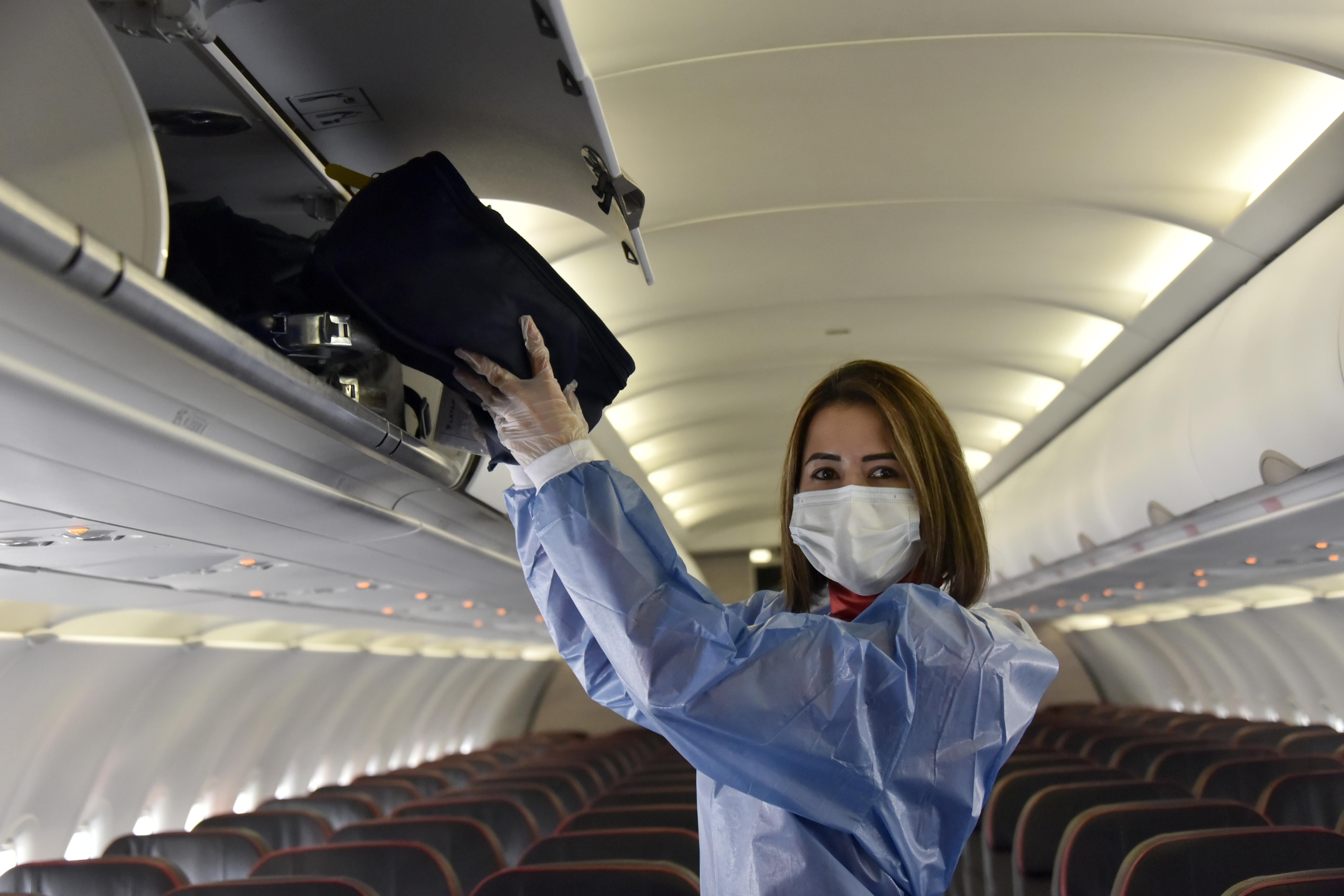 A woman wearing protective equipment, including a mask, stands in the aisle of an empty plane as she puts a bag into an overhead compartment.