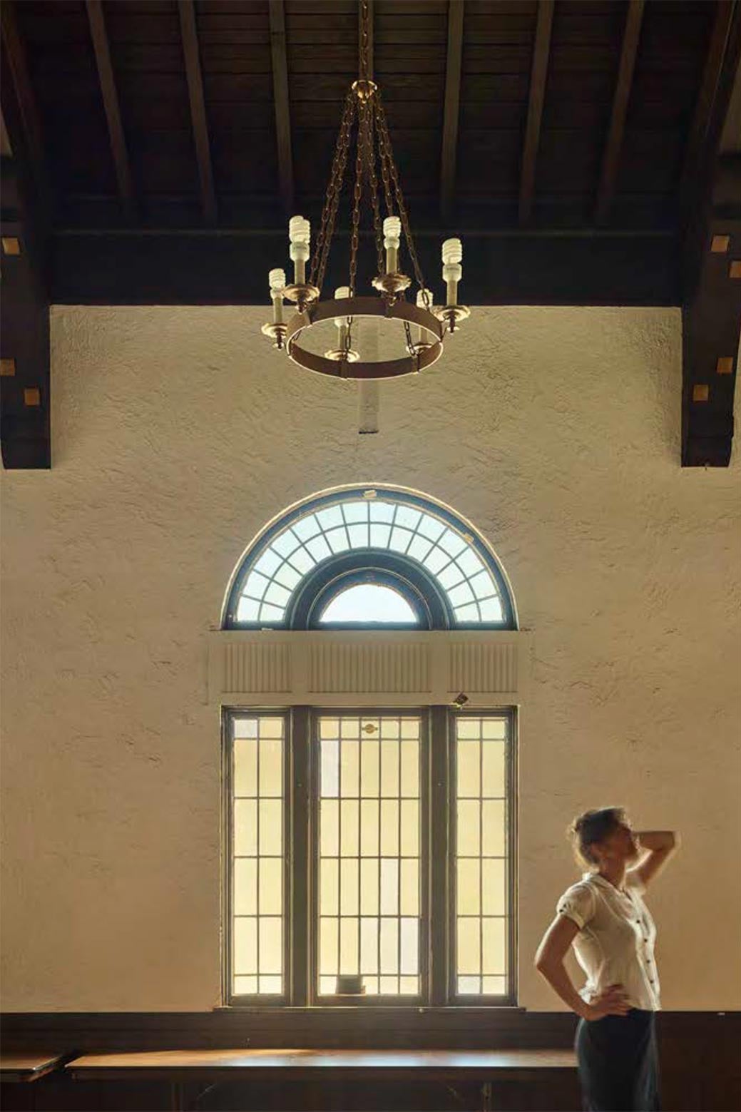 A woman holds her hand to her head and looks up at the room while standing beside a large window and below a chandelier.