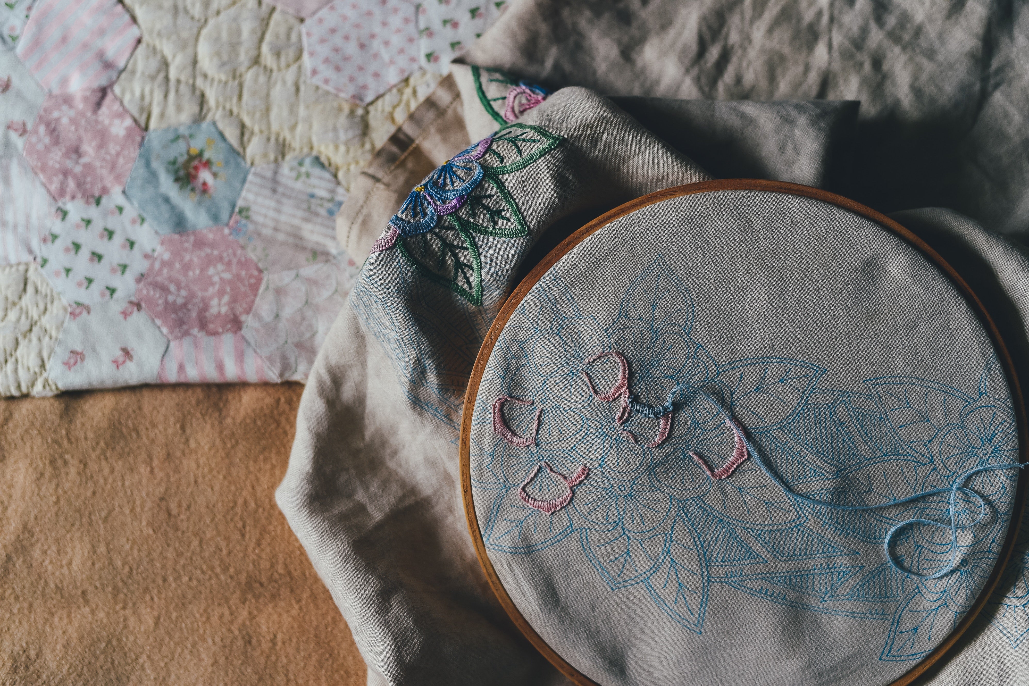 The beginning of an embroidered design lies inside a wooden hoop. A quilt lies in the background.