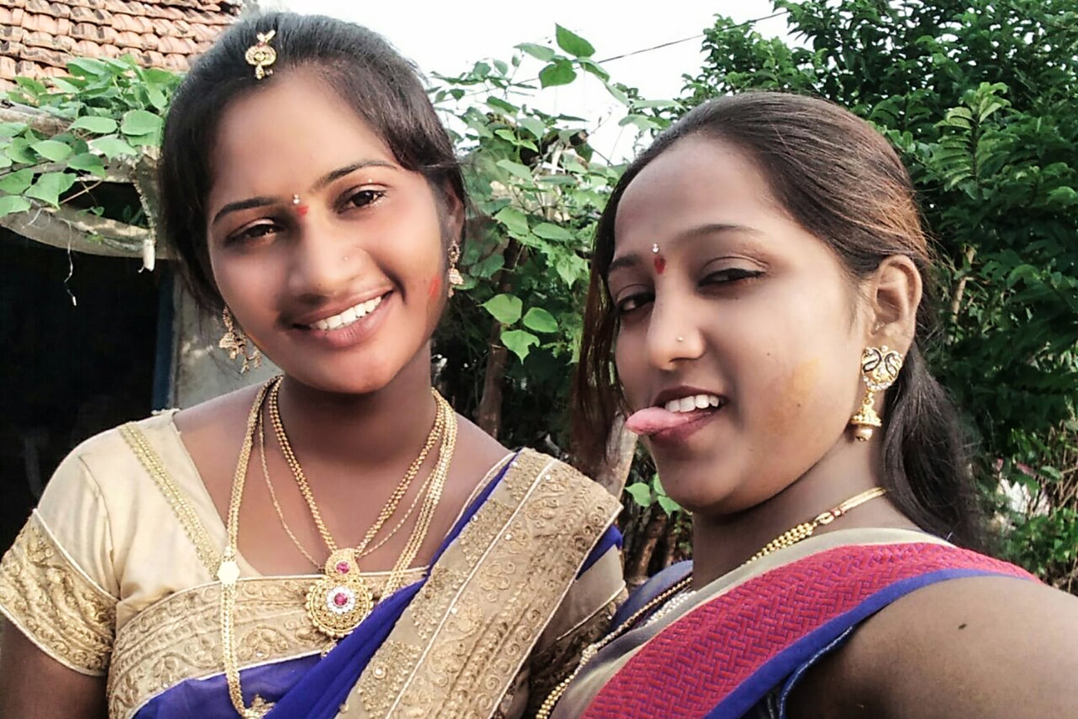 Preethi smiles into the camera, Reshma sticks her tongue out. The women have their arms around each other, each wearing saris and gold jewelry, with trees and a roof in the background