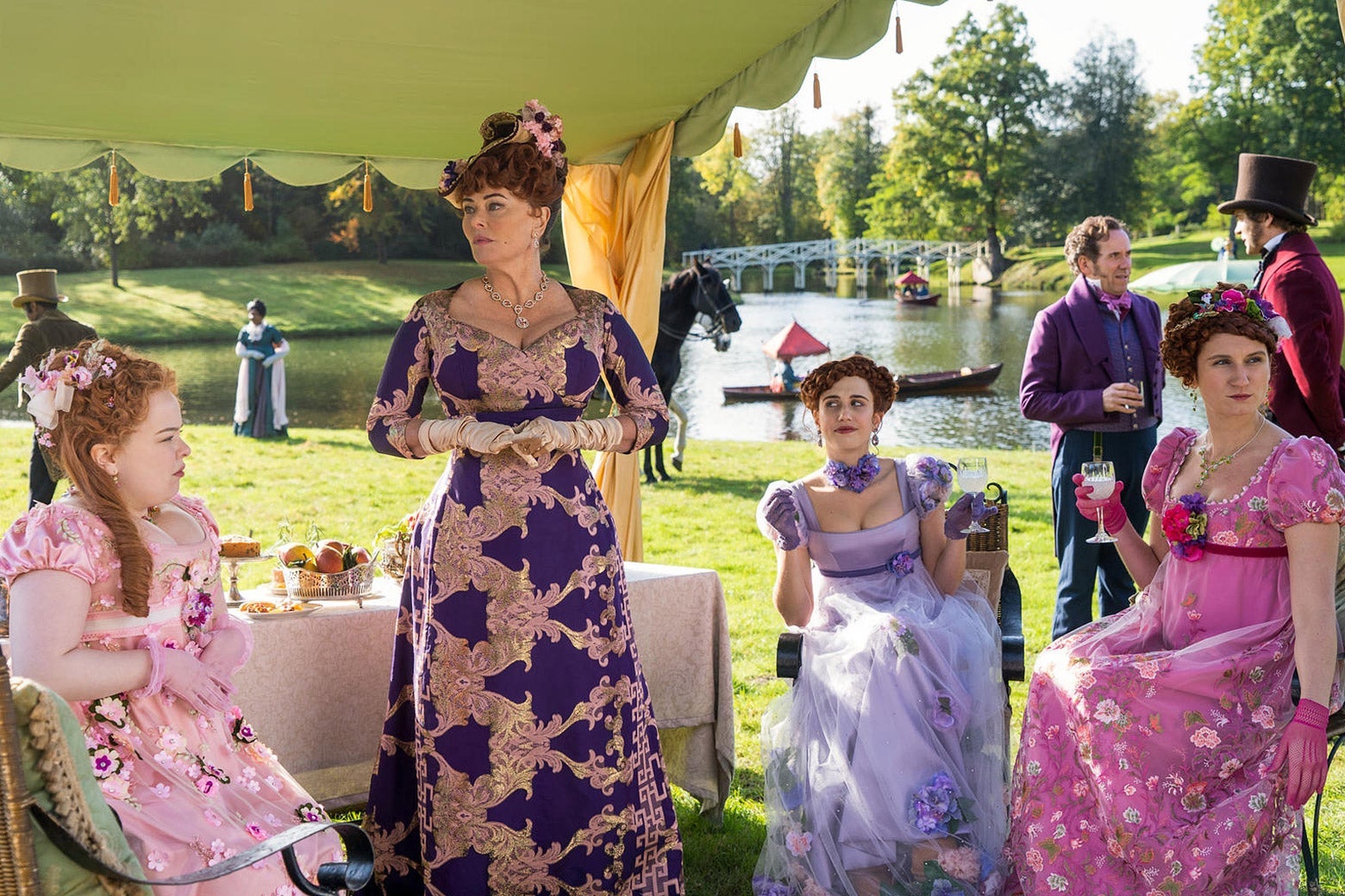 A garden party scene. From left to right: Nicola Coughlan as Penelope Featherington wears a light pink gown with flowers; Polly Walker as Lady Portia Featherington wears a deep purple gown with a bold repeating pattern; Harriet Cains as Philippa Featherington wears a satiny lavender gown with a floral choker and a sheath layered skirt; Bessie Carter as Prudence Featherington wears a bright pink gown with flowers.