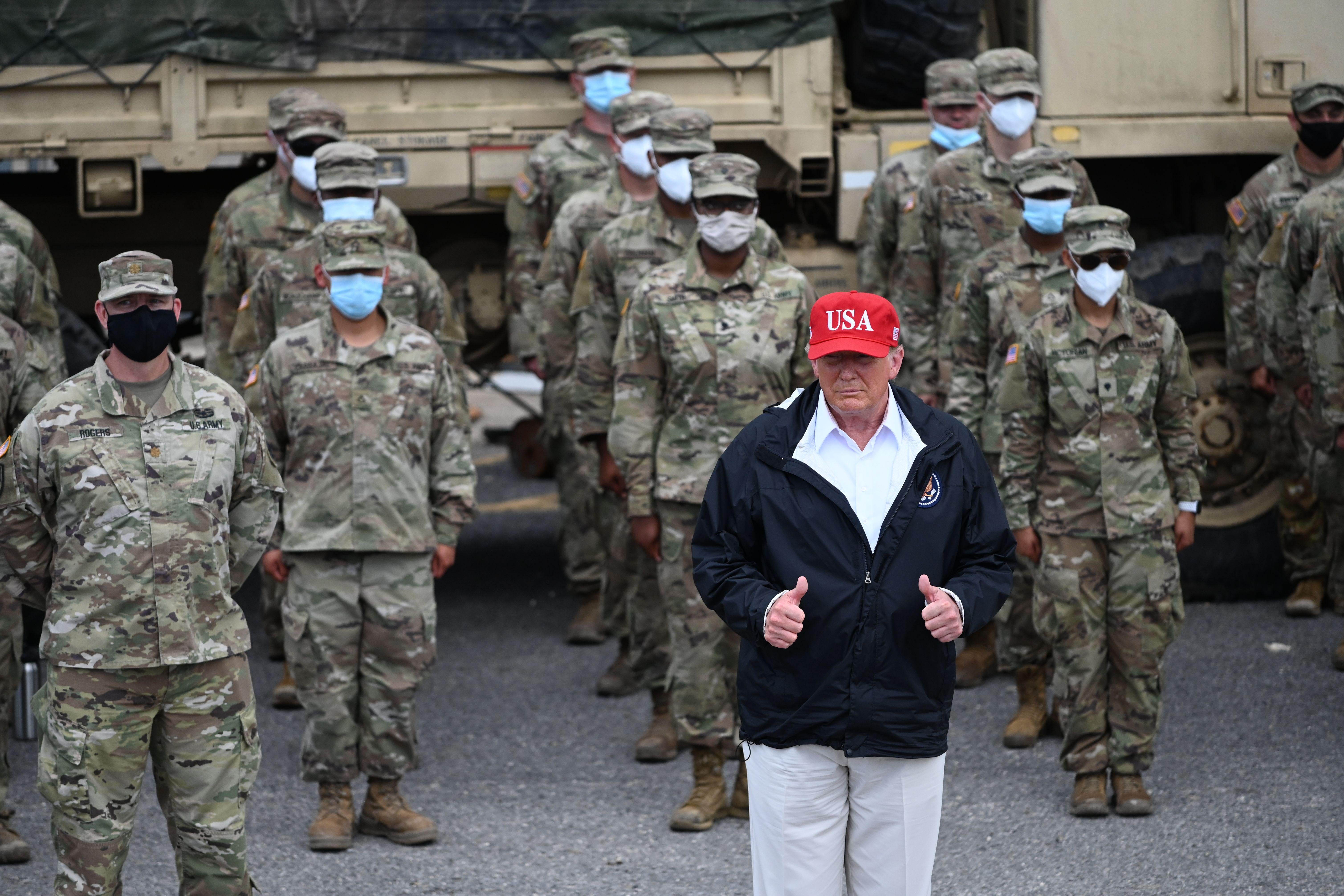 Donald Trump, in a blue jacket and red USA cap, stands in front of rows of National Guardsmen wearing camouflage, sunglasses, and masks. Trump is giving two thumbs up.