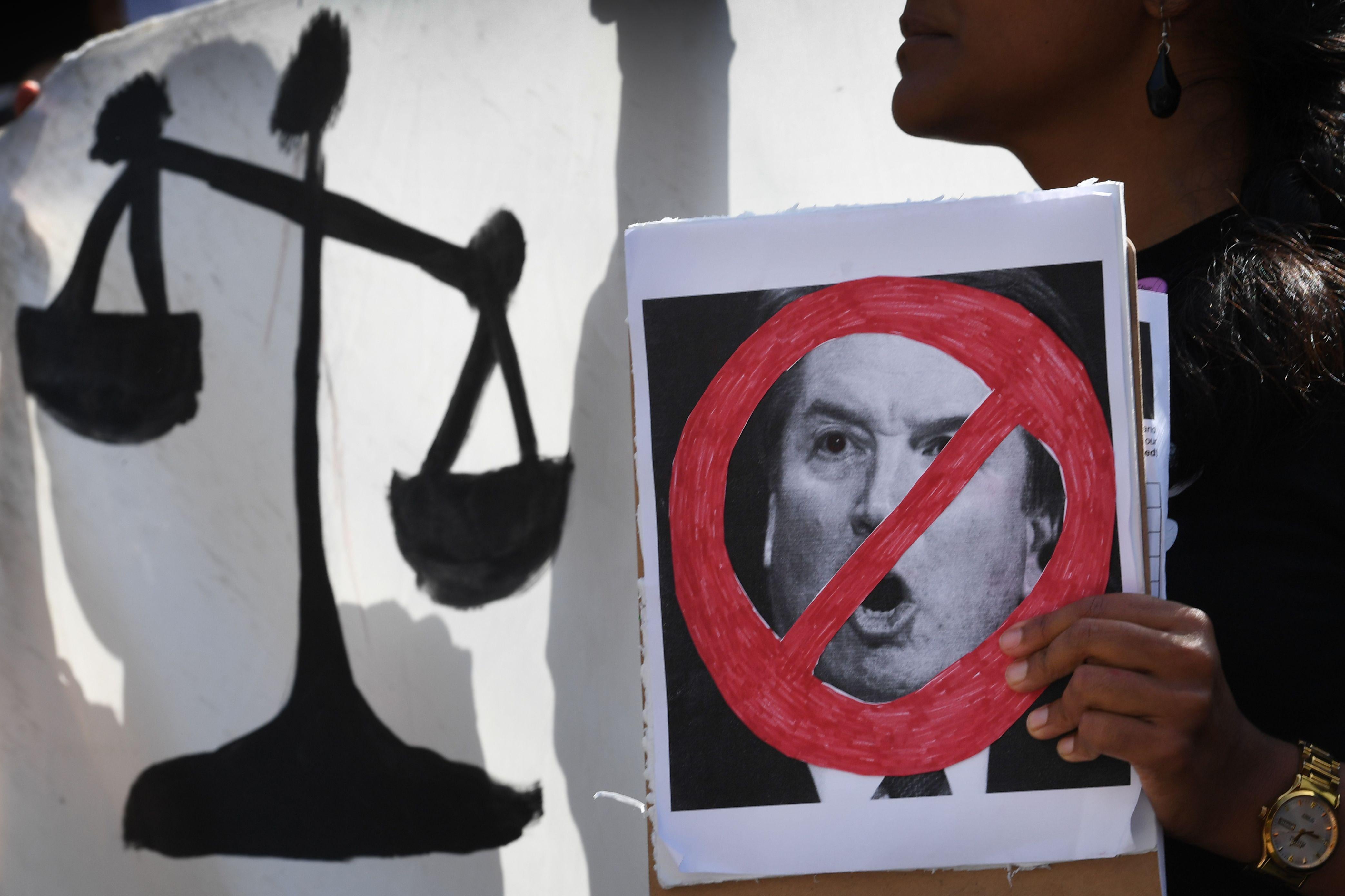 A woman holding a poster showing Kavanaugh's face crossed out, in front of a poster showing the scales of justice.