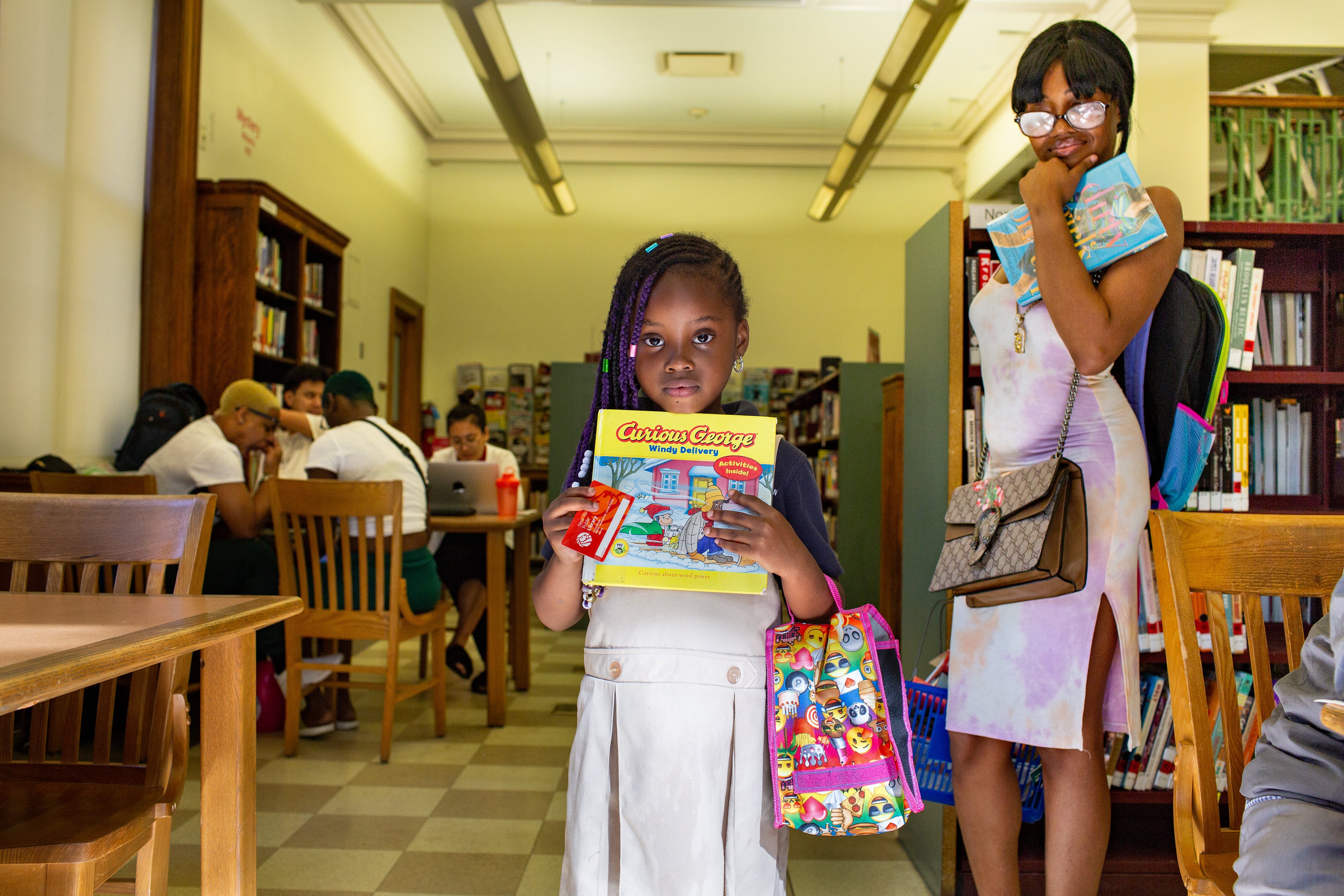 A girl holds a Curious George book and her library card proudly as a young woman looks on. A group of teens hang out at a table in the background.