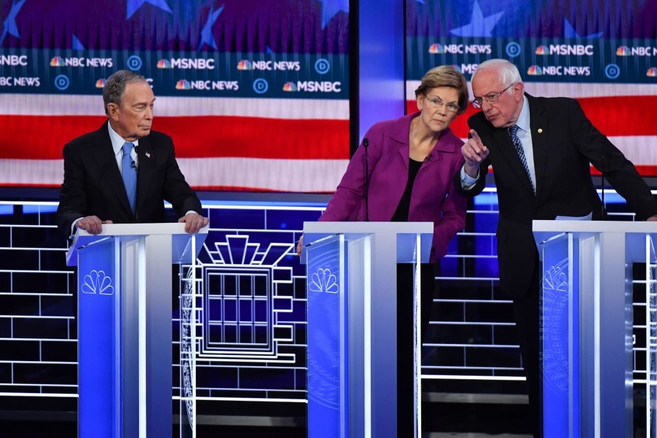 Bloomberg, standing at his lectern, looks to his left with pursed lips while Warren and Sanders lean toward each other.