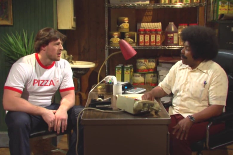 JJ Watt, wearing a white shirt that says "Pizza" in red letters, stares across a desk at Kenan Thompson, in an afro wig. They are in a 1980s industrial kitchen.