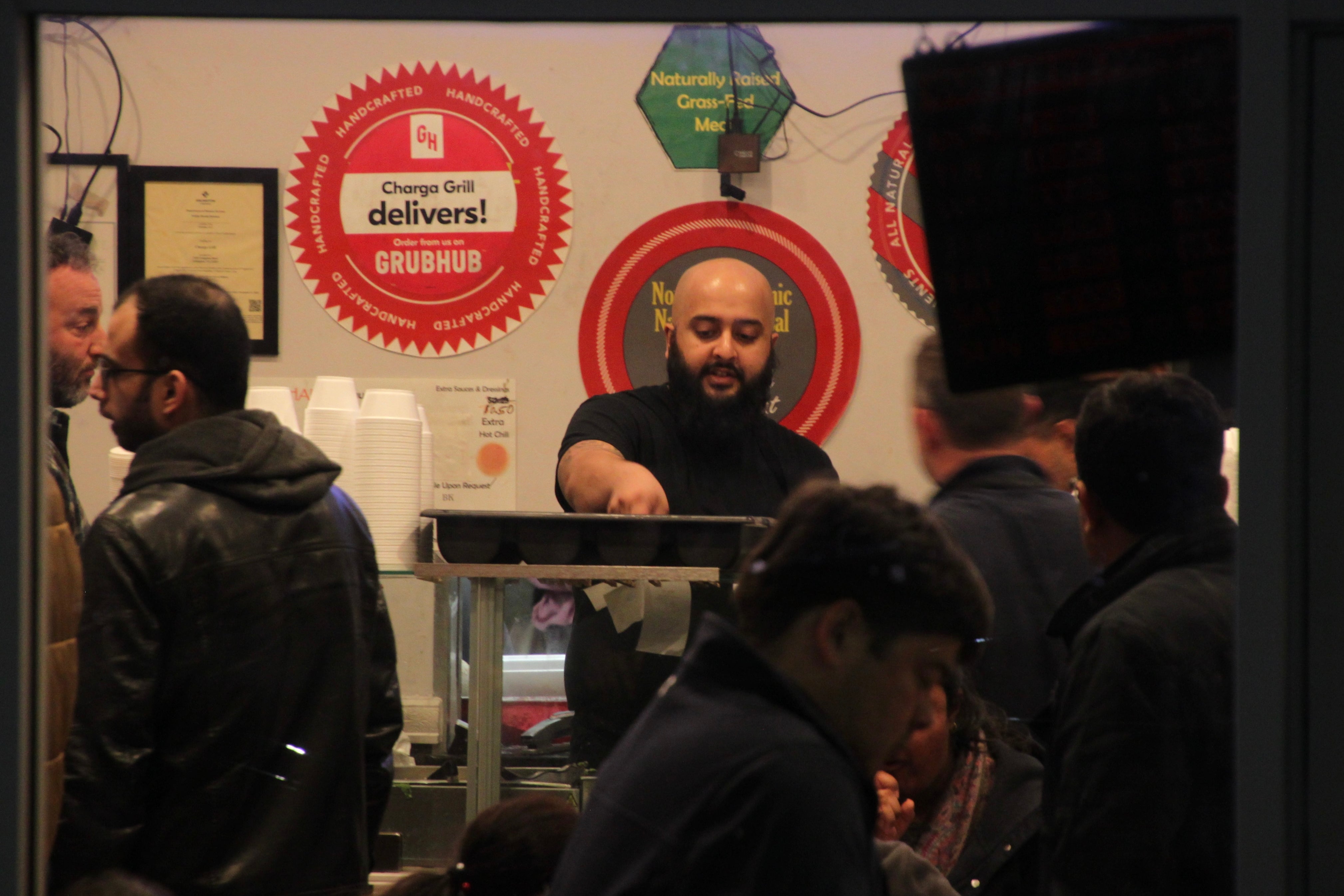 A bald man with a beard stands behind the counter in a packed restaurant.
