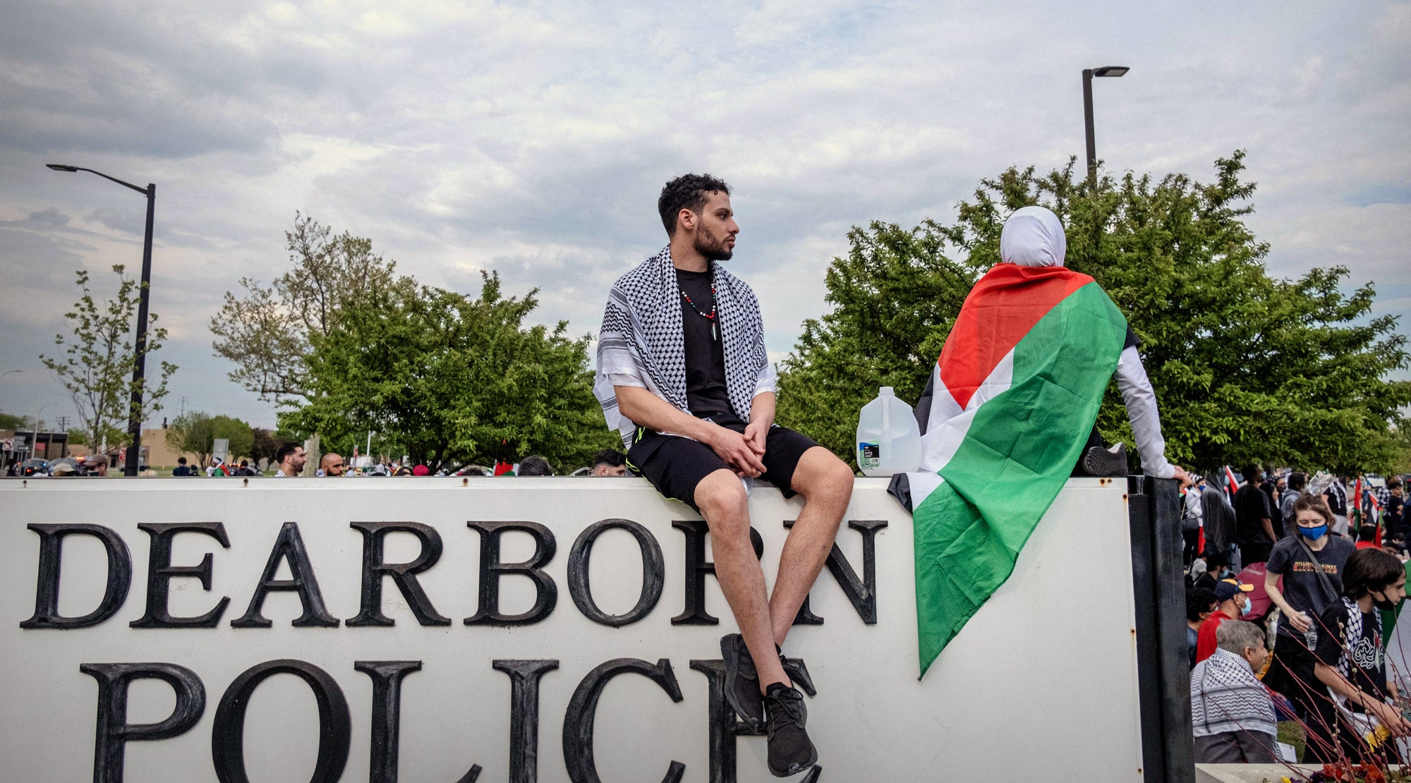 Two people, one draped in a Palestine flag, sit atop a sign that says "DEARBORN POLICE," with a crowd gathered in the grass behind it.