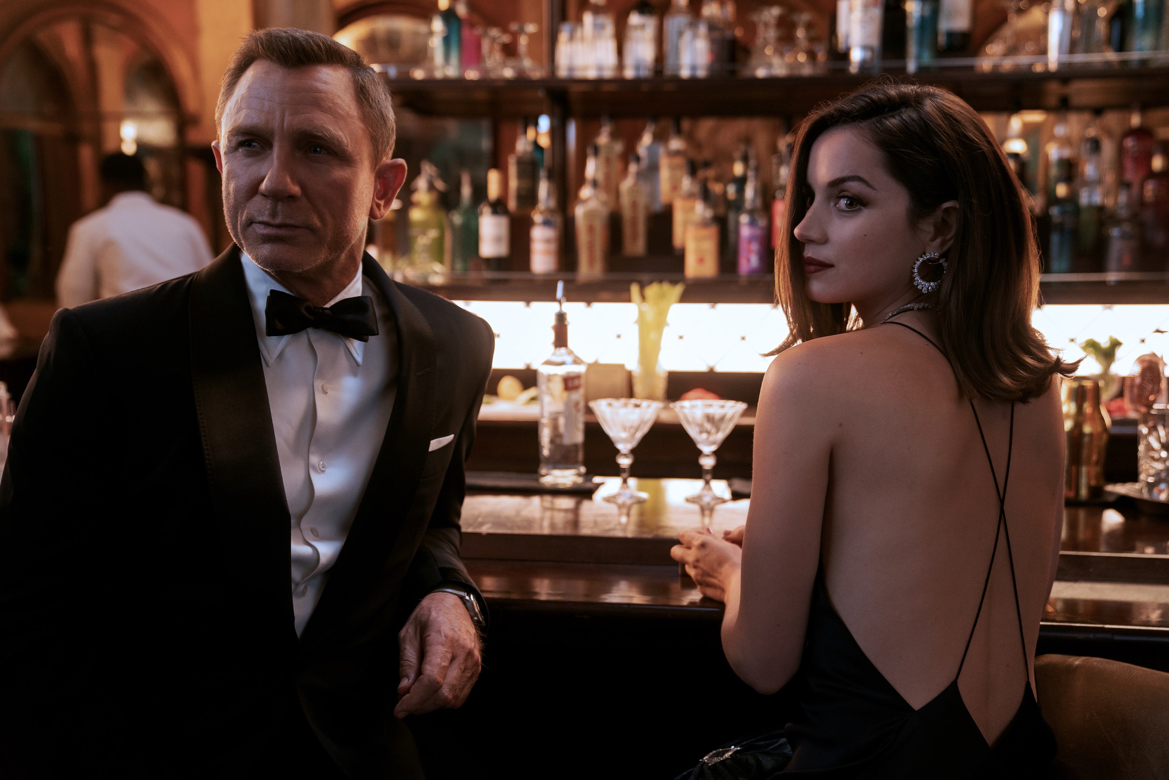 On the left, a blond man wears a tuxedo and looks to his right. He leans on a bar. Next to him is a woman with long brown hair and a strappy black dress, who holds a glass and sits at the bar. She looks in the same direction. 