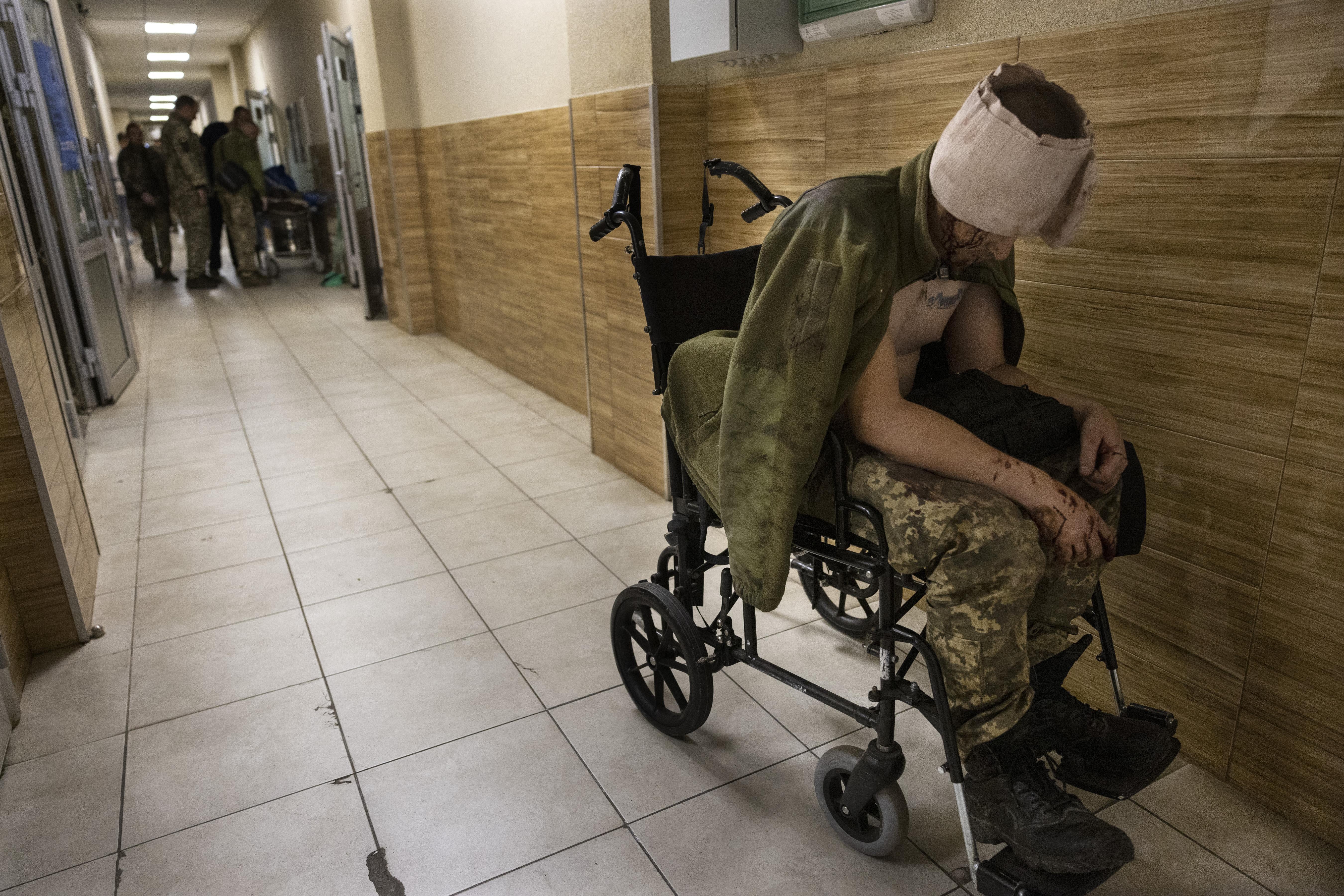 A soldier leans over in a wheelchair. His head is wrapped in bandages.