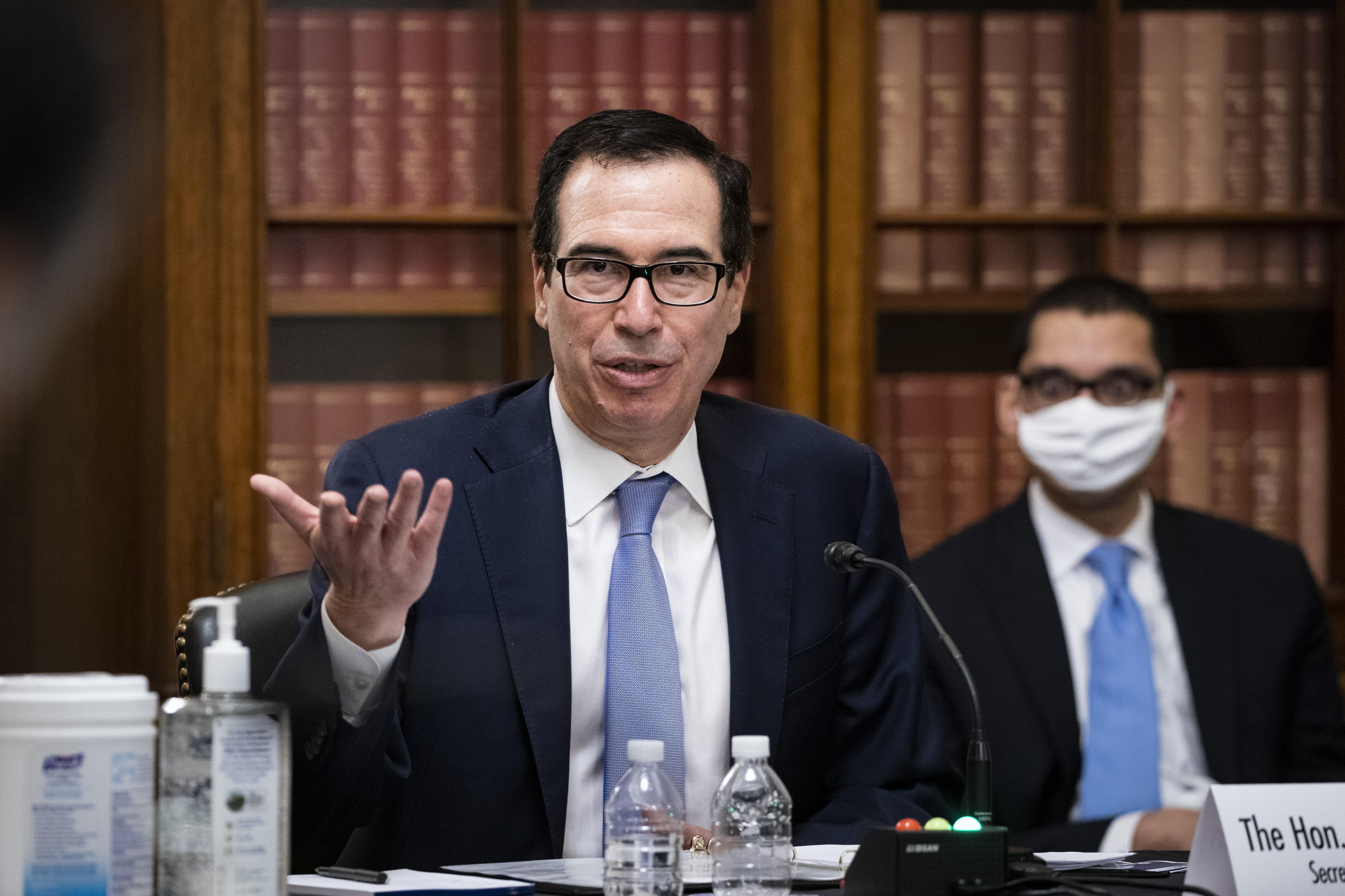 Steven Mnuchin sits at a table and raises his hand while speaking. A man sits behind him, wearing a face mask.