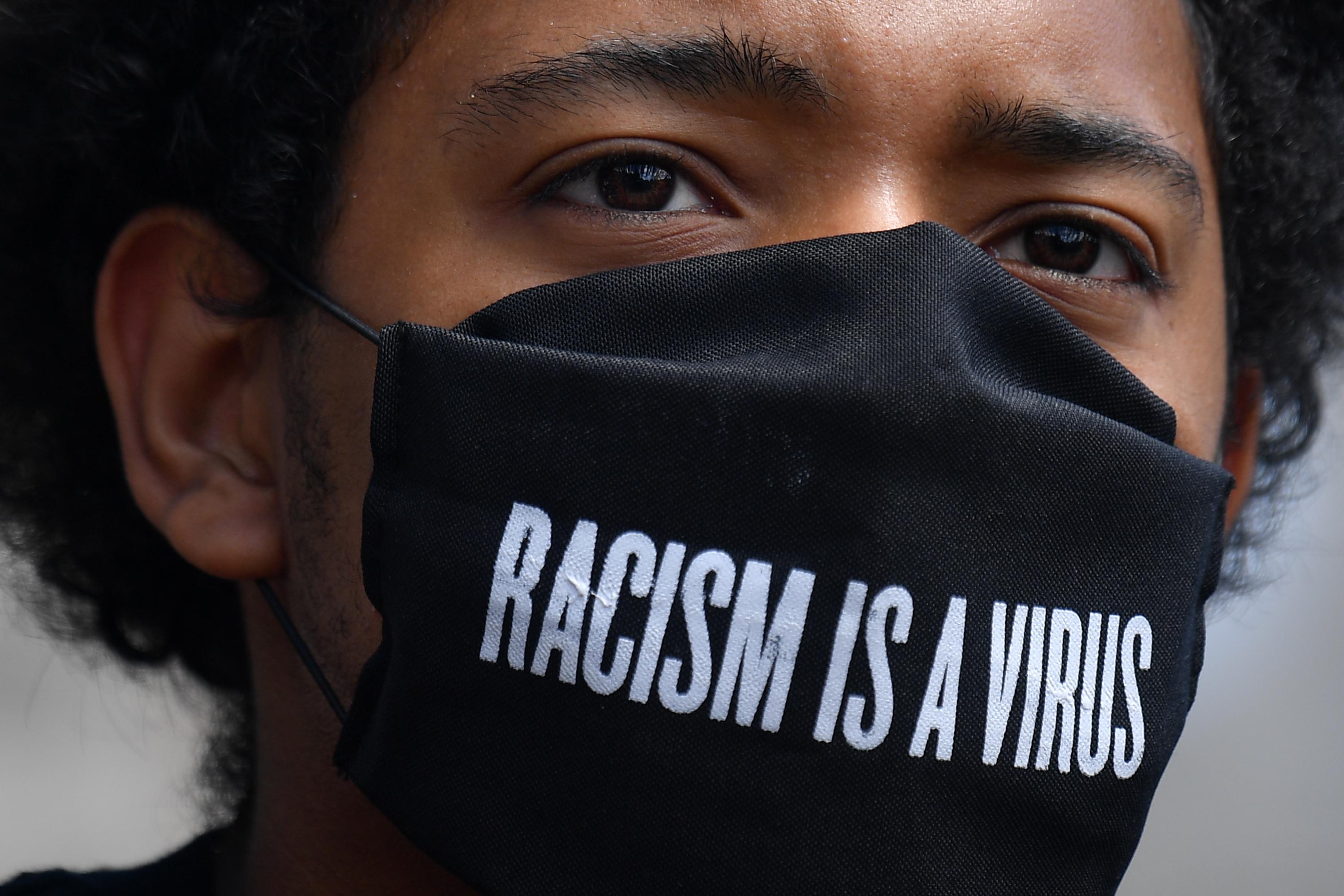 A protester wears a protective face covering with the slogan "Racism is a Virus" written on the fabric as he listens to speeches at a gathering in support of the Black Lives Matter movement at Marble Arch in London on July 5, 2020.