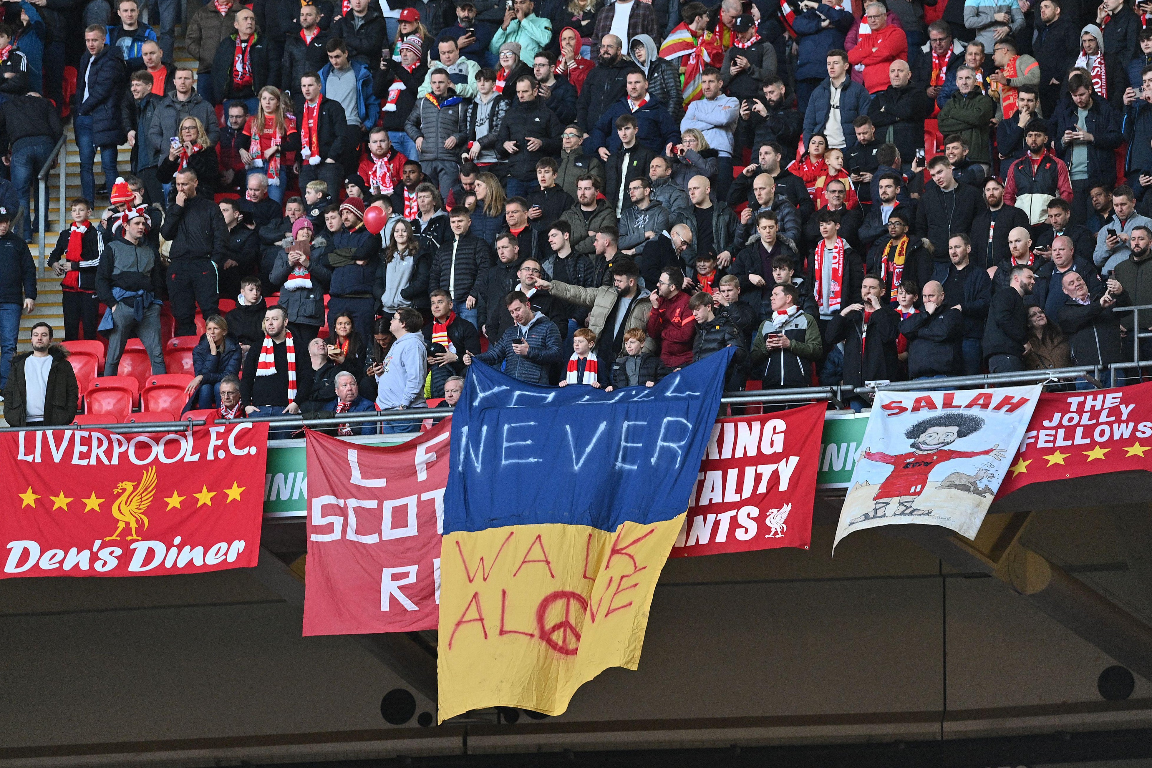 Liverpool supporters hold up a Ukrainian flag with the team’s motto “You’ll Never Walk Alone” written on it, a peace sign illustrated in the "o" of "Alone," amid general signs supporting Liverpool FC and team star Mo Salah.