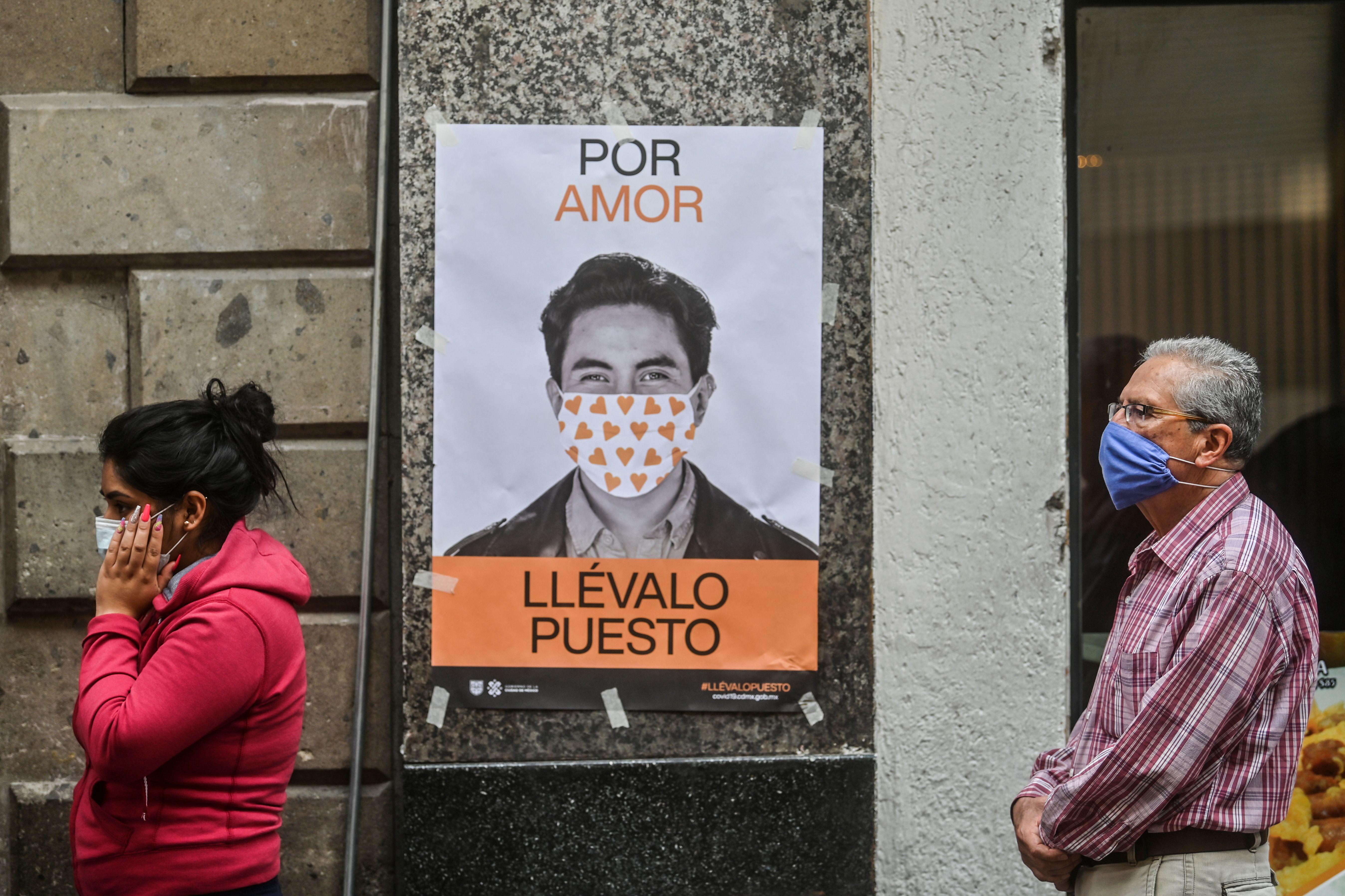 People wearing face masks walk past a poster with a photo of a man in a face mask and the caption "Por Amor, Llévalo Puesto," or "For Love, Use It." 