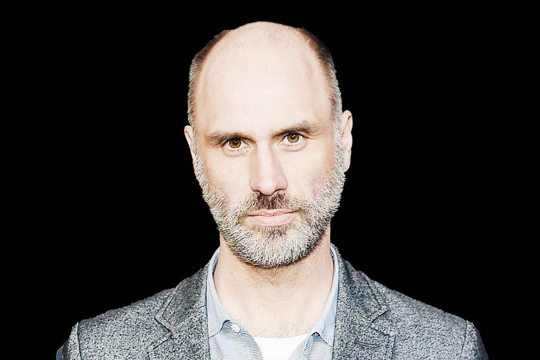 A balding man with a beard stares straight ahead at the camera.