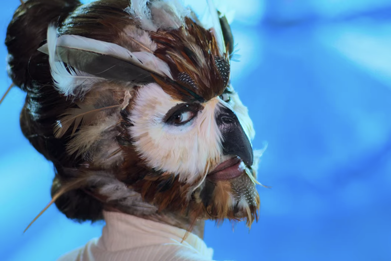 Woman in a white turtleneck and an owl mask.