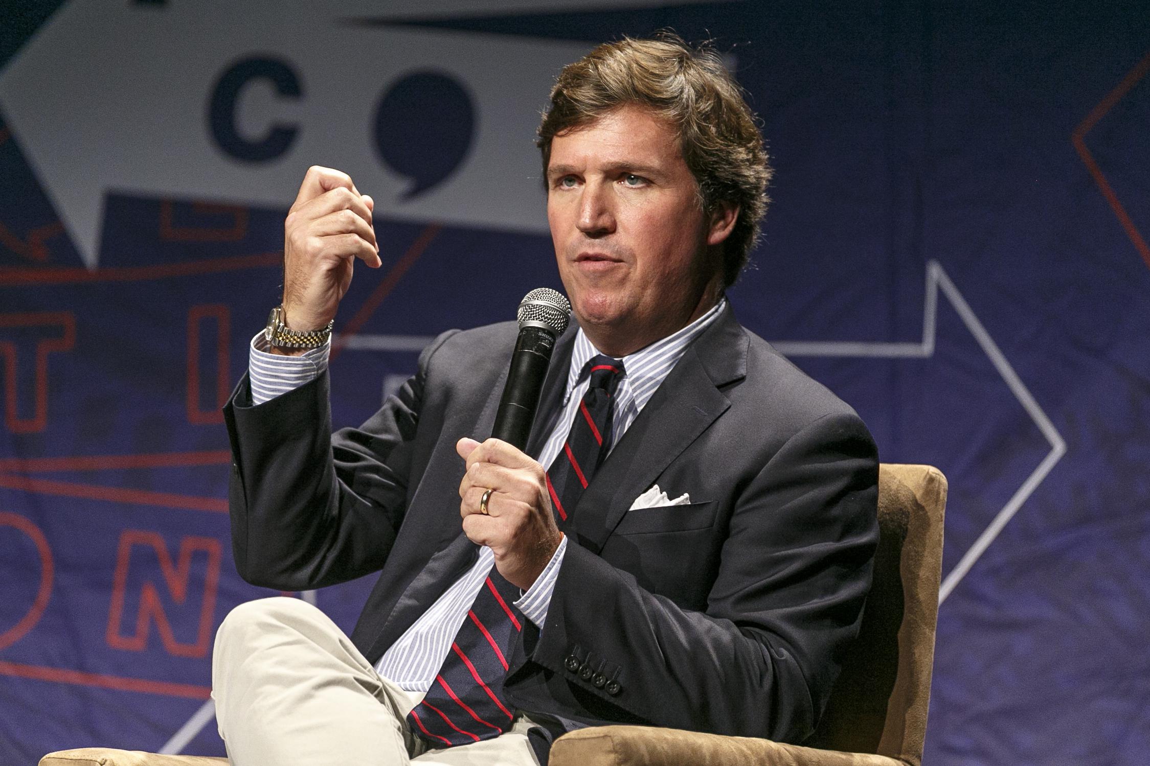 Carlson sits casually in a chair on a stage and speaks into a microphone while gesturing with his hand.