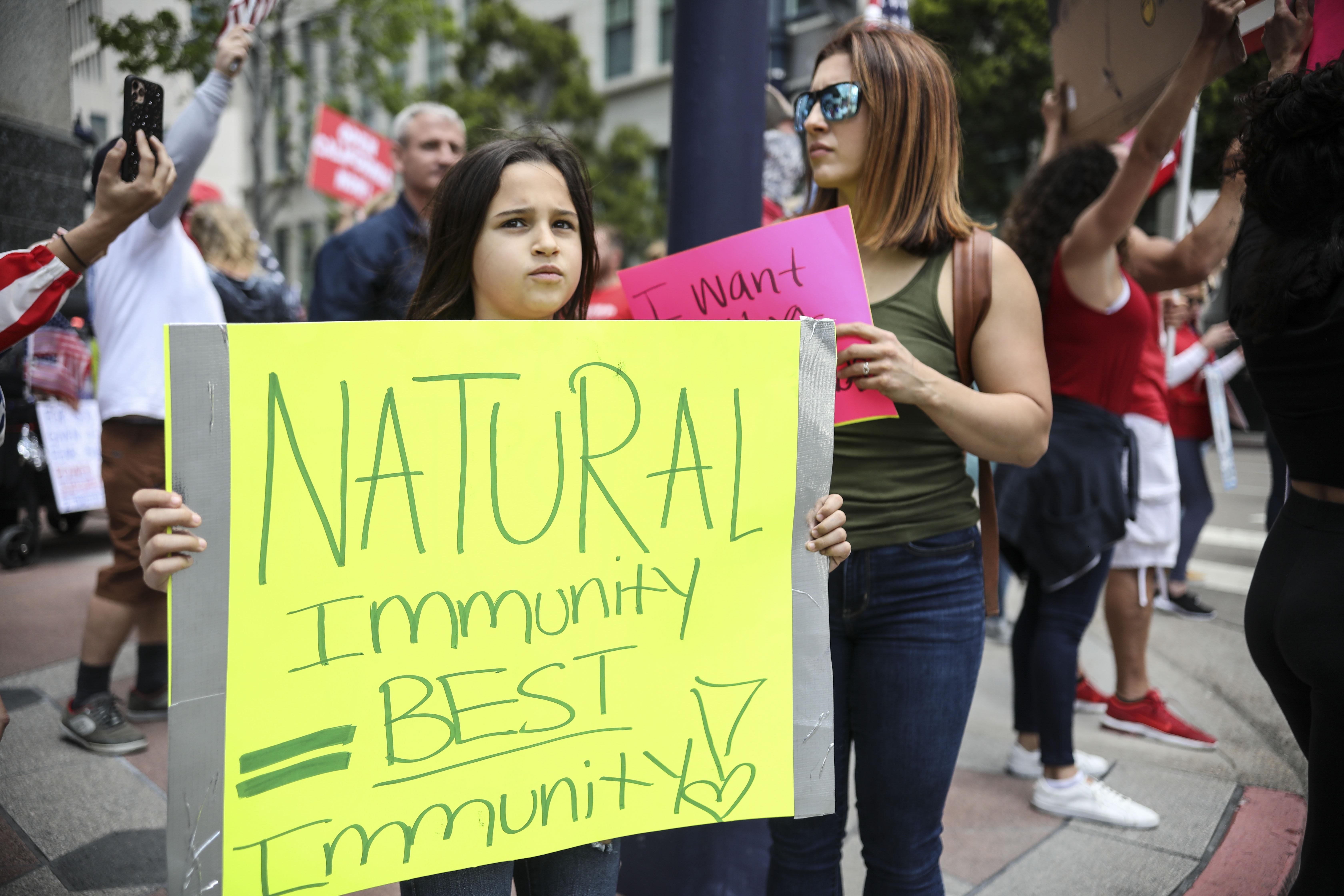 Several protesters on the street, one of them a child holding a sign that says "Natural Immunity = Best Immunity"