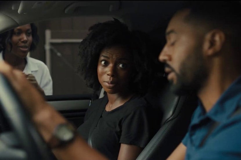 A woman and a man sit in a car, another woman looks through the passenger car window