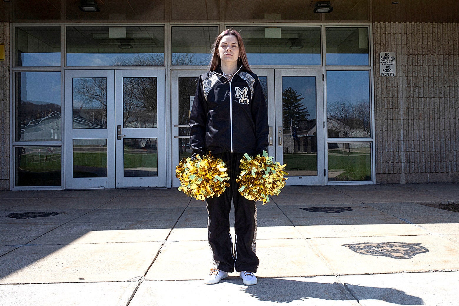 A woman in a track suit holds pompoms in front of a school.