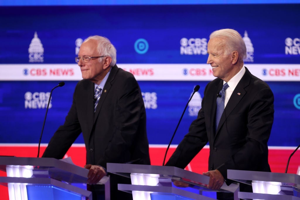 Sanders and Biden stand at adjacent lecterns while smiling.