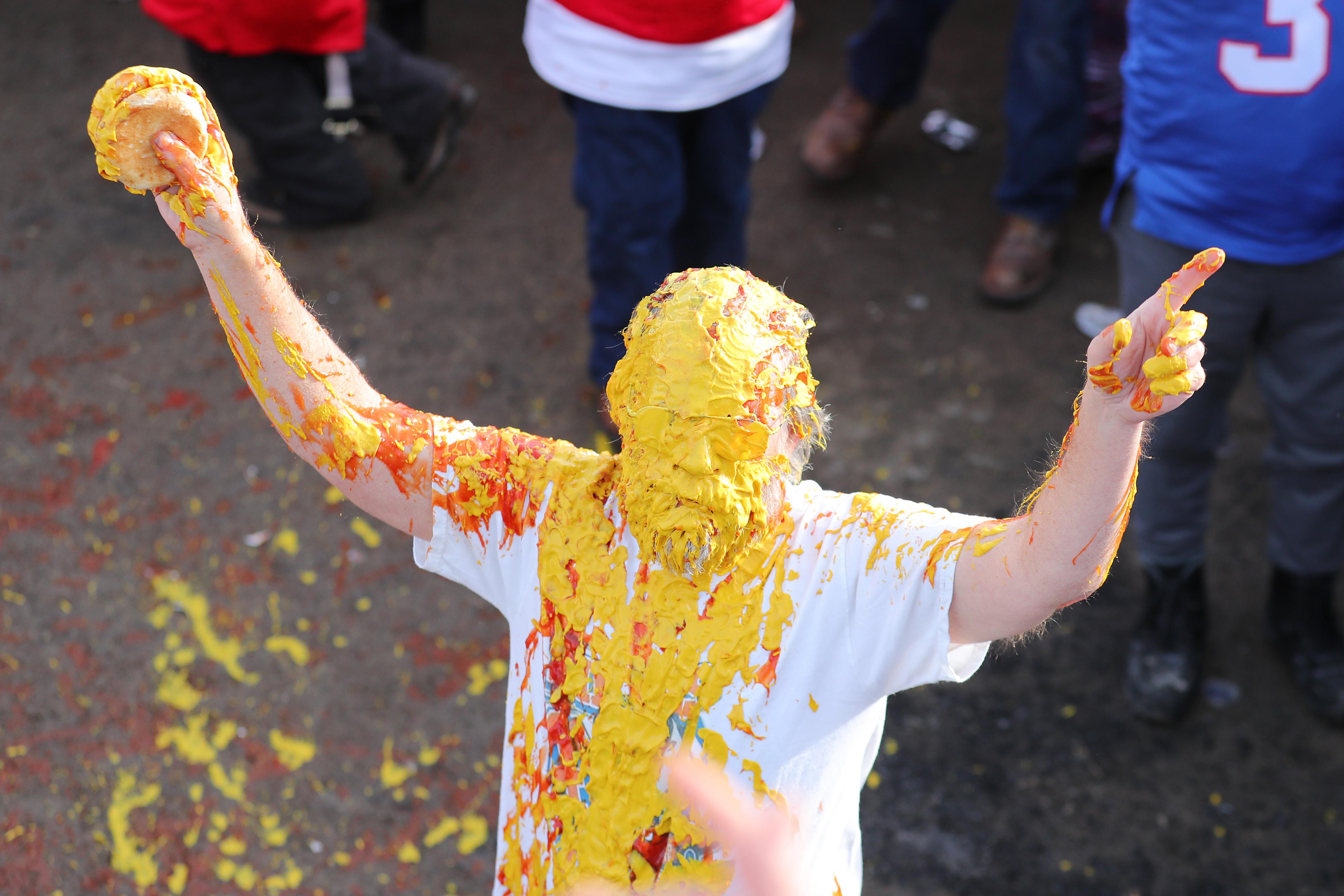 Ken "Pinto Ron" Johnson, holding a burger in his right hand hand and raising his left index finger, is covered in a thick layer of yellow mustard and some ketchup all over his face and down his shirt.