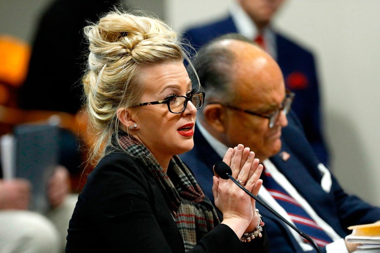 Melissa Carone at a table speaking into a microphone next to Rudy Giuliani.