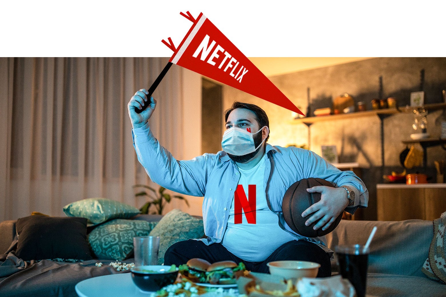 A fan of Netflix sits on his couch, wearing his Netflix jersey, waving his Netflix pennant, wearing a surgical mask