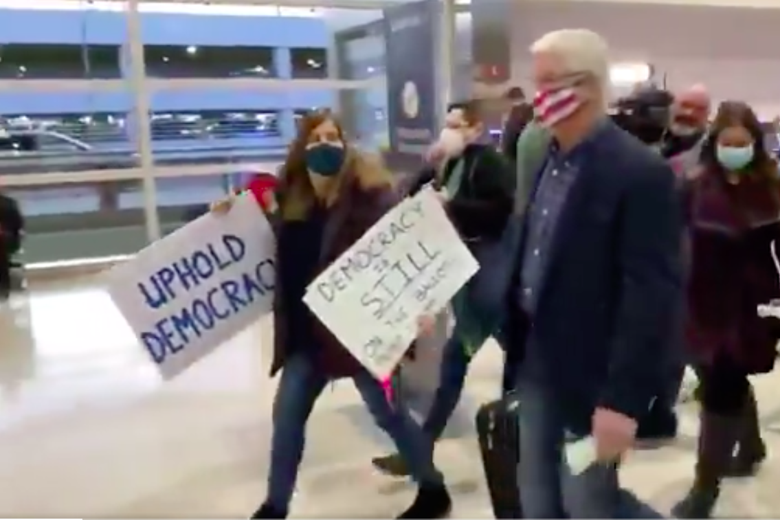 Michigan Senate Majority Leader Mike Shirkey walks through an airport with his rolling suitcase, surrounded by protesters holding signs. One sign says "Uphold Democracy."