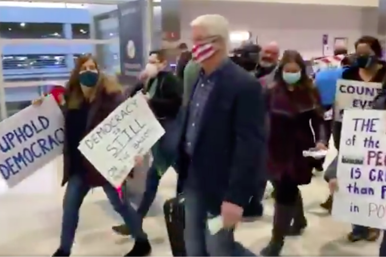Michigan Senate Majority Leader Mike Shirkey walks through an airport with his rolling suitcase, surrounded by protesters holding signs. One sign says "Uphold Democracy."