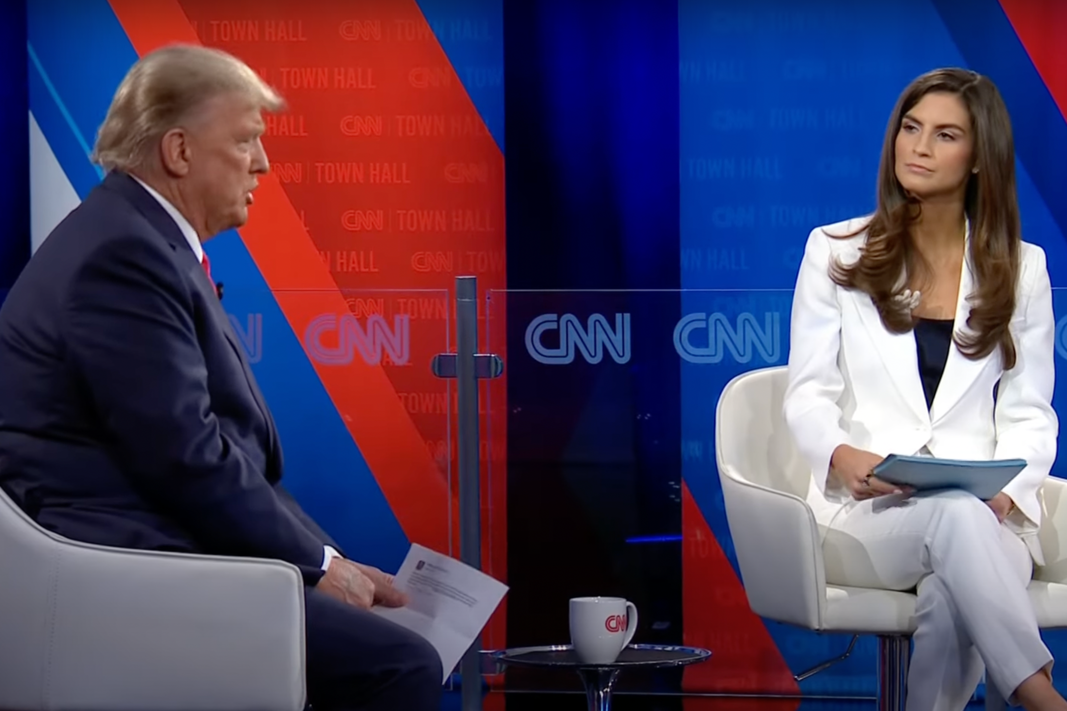 Trump sits in a patriotically appointed auditorium, speaking while holding a piece of paper, while Kaitlan Collins listens.