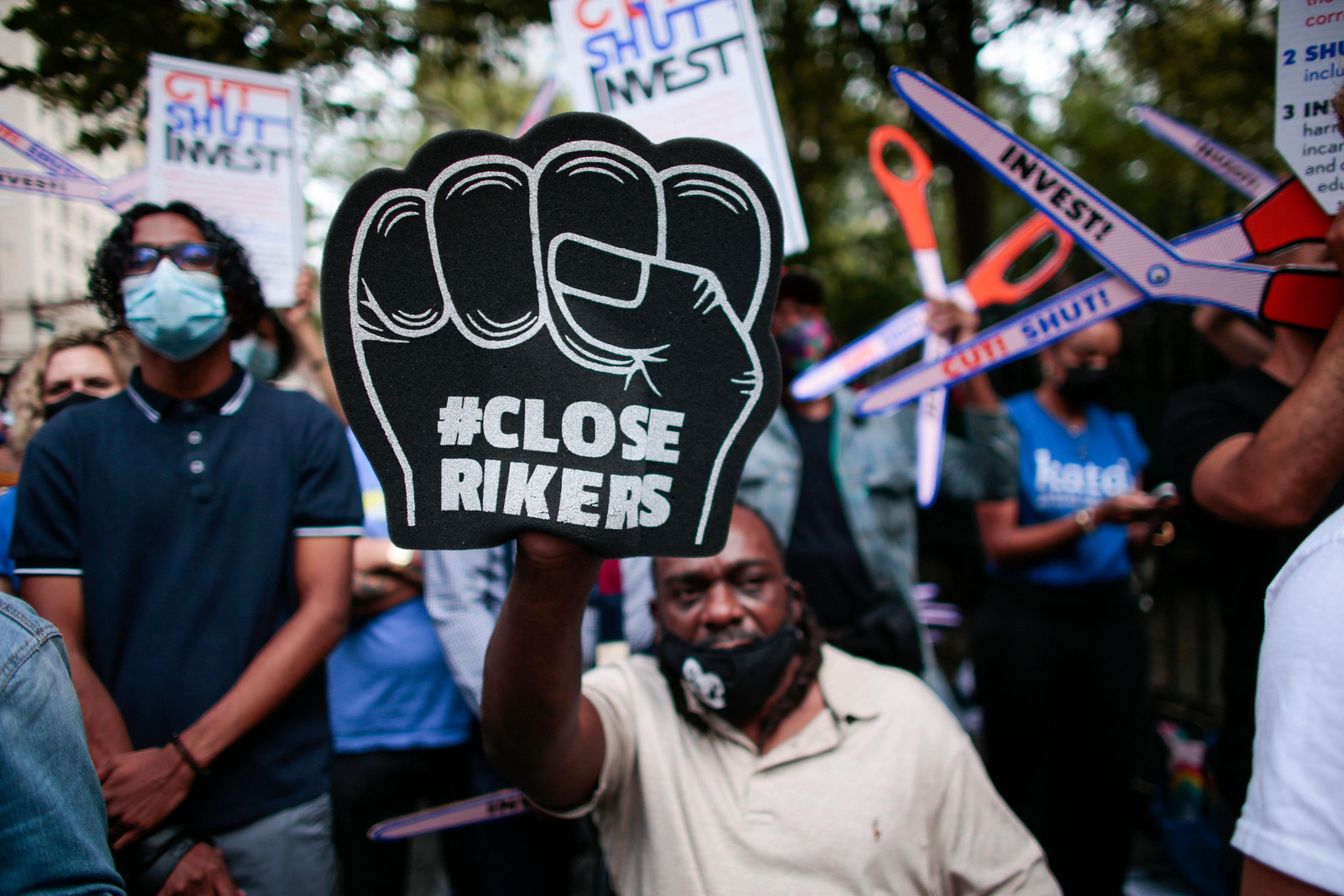 A Black man holds up a black foam fist that says, "#CloseRikers."