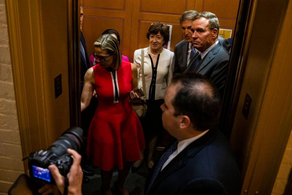 The four senators stand in an elevator as Sinema presses a button.